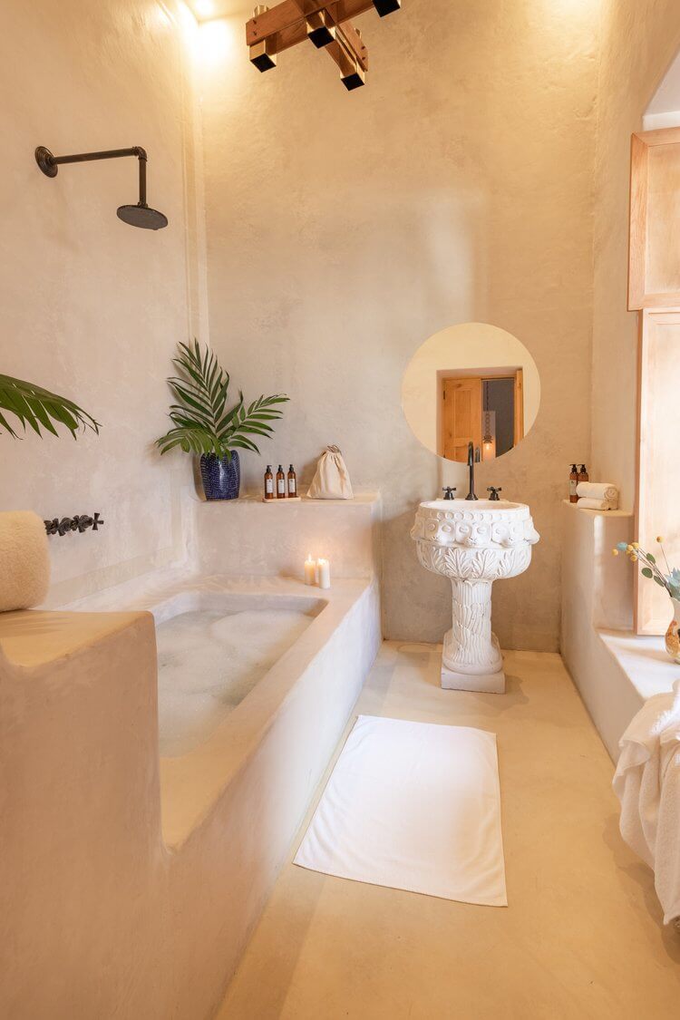 La Valise San Miguel, Mexico — Cream-hued bathroom with sculptural pedestal sink, soaking tub, round mirror, and potted palm under wooden ceiling fixture.