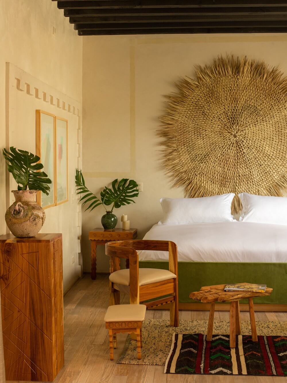 La Valise San Miguel, Mexico — Ochre bedroom with woven sunburst headboard, wooden furniture, monstera plants, and colorful woven textiles on terracotta floors.