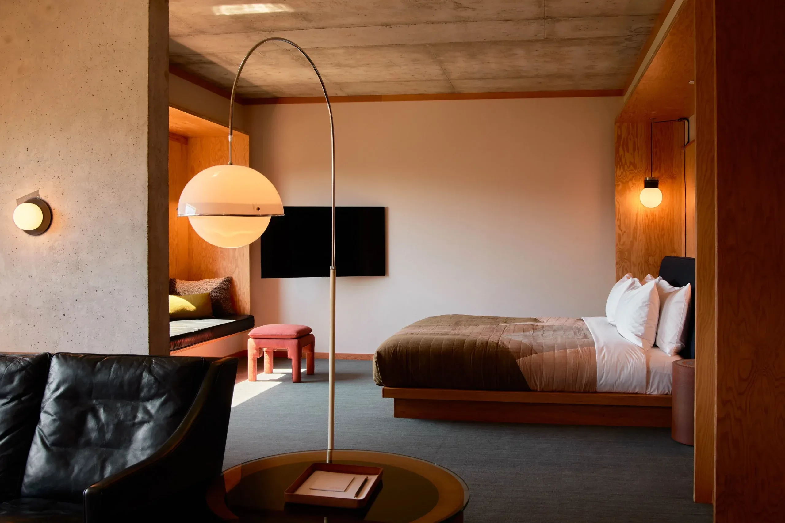 Ace Hotel Toronto, Canada — Minimalist hotel suite with arc floor lamp, leather seating, wooden platform bed, concrete walls, and warm ambient lighting.