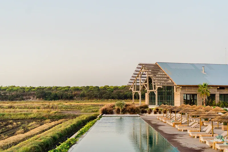 Quinta da Comporta, Portugal — Contemporary farmhouse with peaked roof beside reflecting pool, rural landscape, wooden lounge chairs, natural light, minimalist architecture.