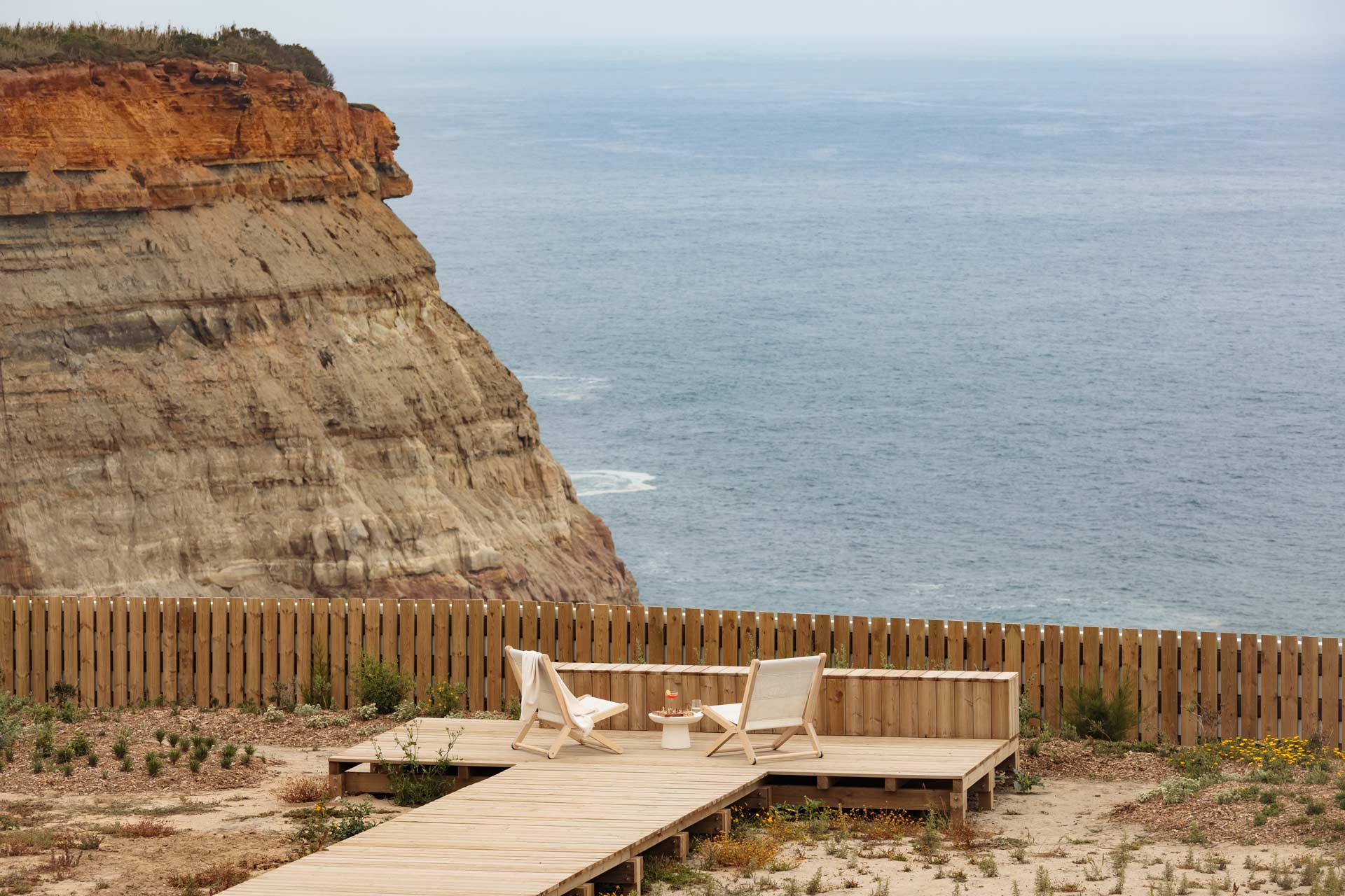 Aethos Ericeira, Portugal — Wooden deck with canvas chairs overlooking ocean cliffs and wooden fence, minimalist coastal design.