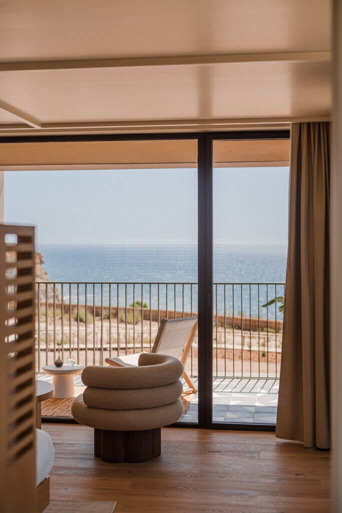 Aethos Ericeira, Portugal — Modern hotel suite with stacked round armchair, wooden floors, floor-to-ceiling glass doors opening to ocean view balcony, minimalist coastal design.