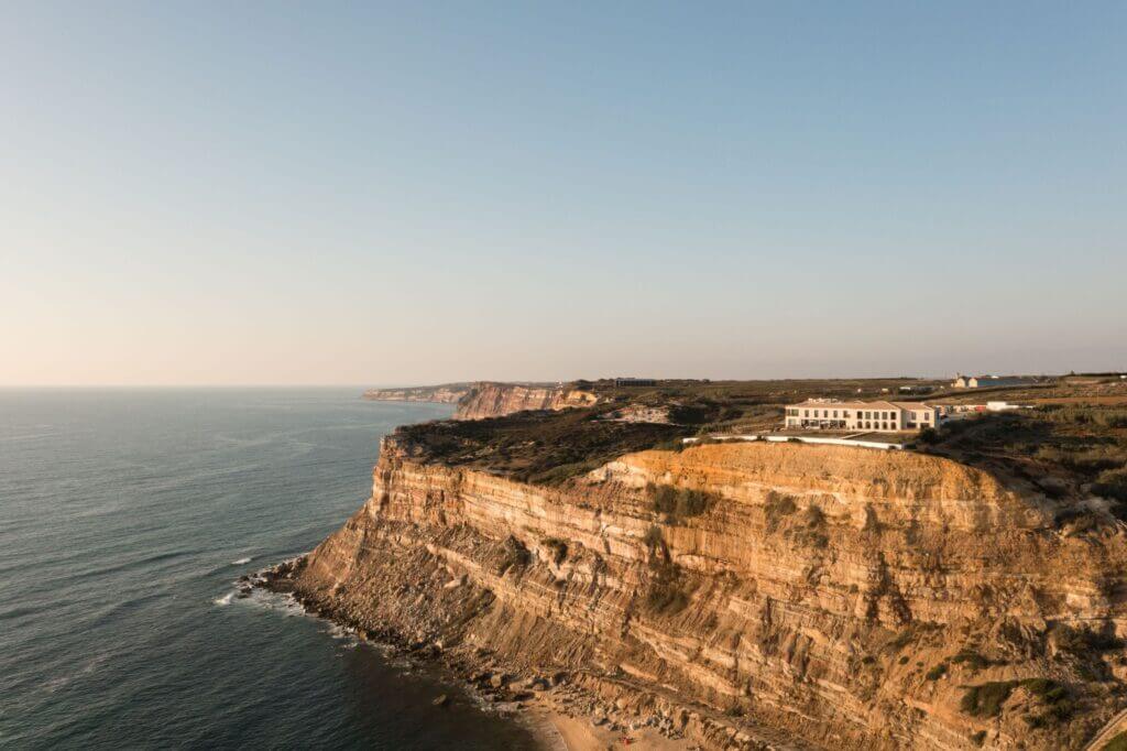 Aethos Ericeira, Portugal — Clifftop luxury hotel with golden stone facade overlooking Atlantic Ocean and dramatic coastal cliffs at golden hour.