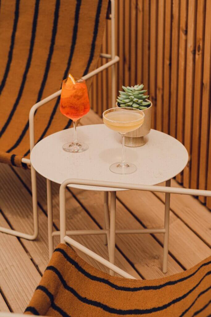 Aethos Ericeira, Portugal — Minimalist white bistro table with aperitif cocktails and succulent plant against warm wood-paneled wall with blue striping.