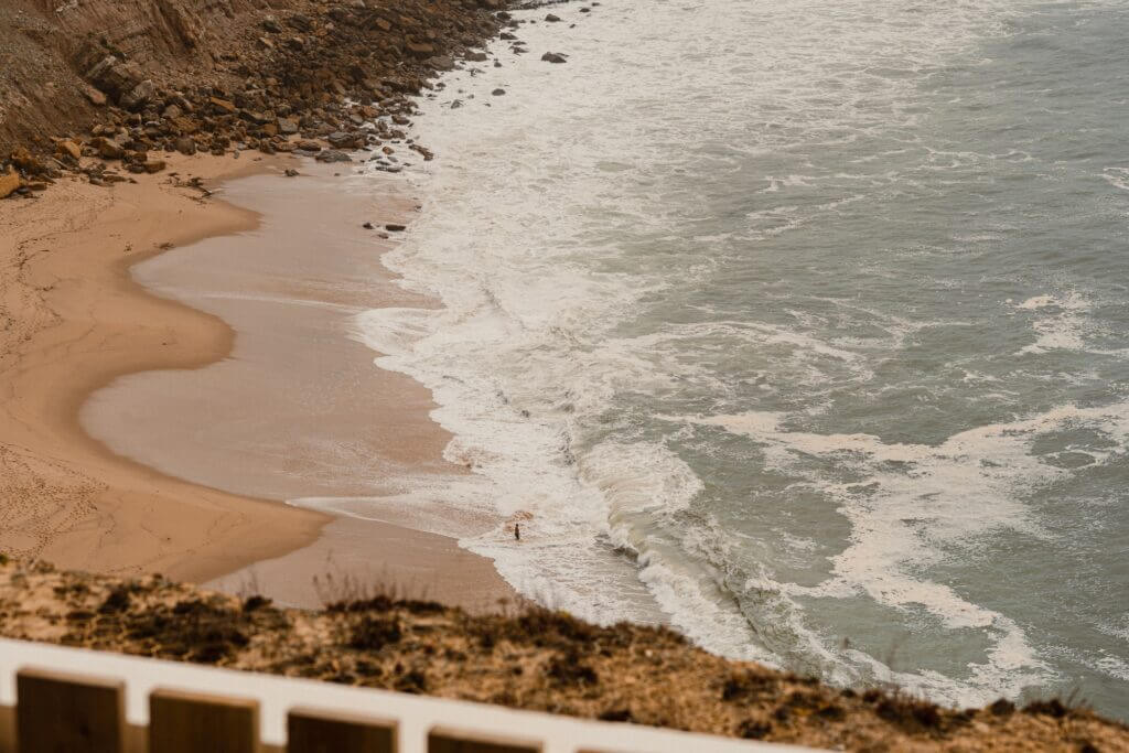 Aethos Ericeira, Portugal — Coastal cliffs overlooking sandy beach with rolling waves and whitewashed surf, aerial view.