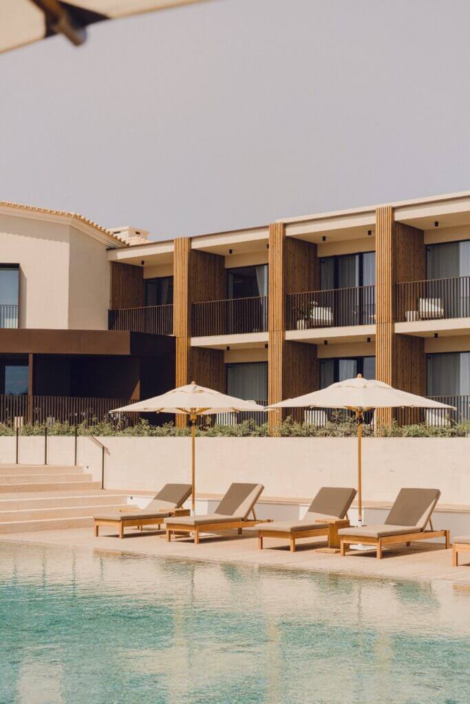 Aethos Ericeira, Portugal — Minimalist poolside lounge with wooden sun loungers, cream umbrellas, and contemporary architecture featuring vertical wood slats and clean horizontal lines.