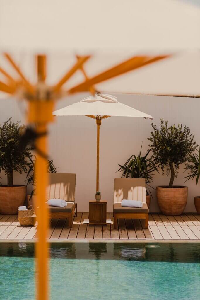 Aethos Ericeira, Portugal — Minimalist poolside lounge with wooden sunbeds, white umbrella, terracotta planters, and soft coastal light at sunset.