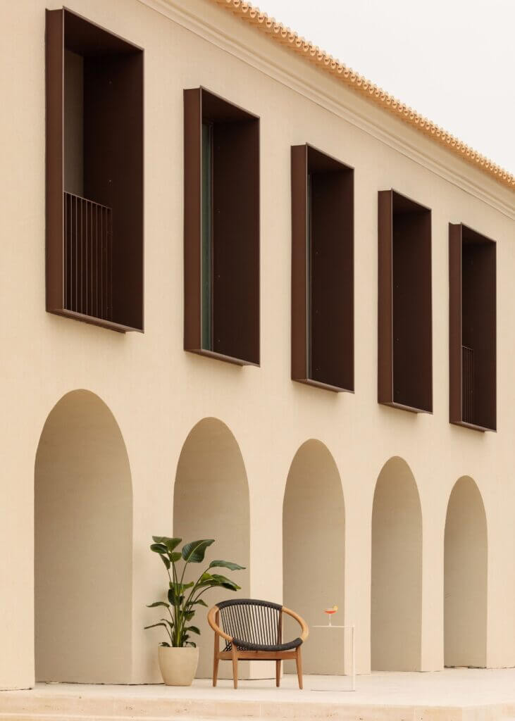 Aethos Ericeira, Portugal — Minimalist courtyard with arched alcoves, wooden chair, potted plant, dark rectangular windows, terracotta roof edge, warm neutral tones.
