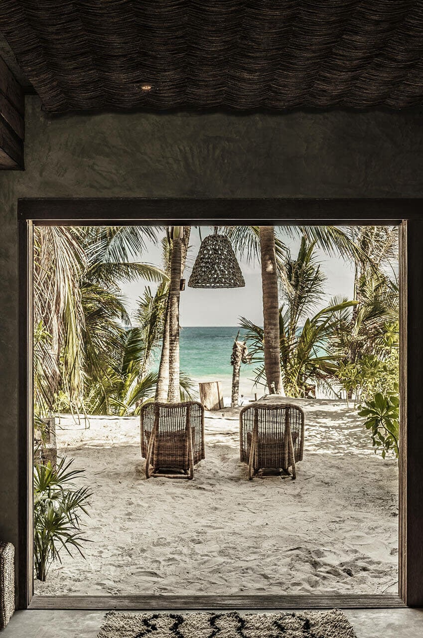 Be Tulum, Mexico — Open pavilion with woven pendant lamp, rattan loungers, palm trees, and turquoise ocean view from sandy beach at sunrise.