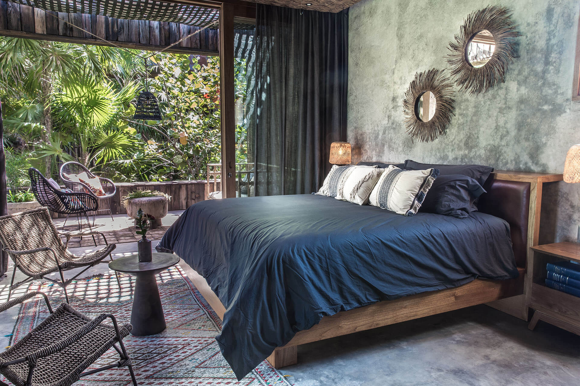Be Tulum, Mexico — Modernist bedroom with wooden platform bed, navy linens, textured concrete walls, decorative sunburst mirrors, tropical garden views through floor-to-ceiling glass, woven lounge chairs on terrace.