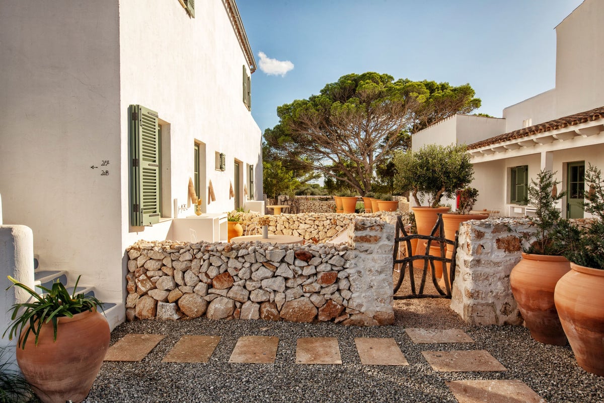 Menorca Experimental, Balearic Islands — Whitewashed courtyard with stone walls, terracotta pots, geometric paving, and Mediterranean pine tree under blue sky.