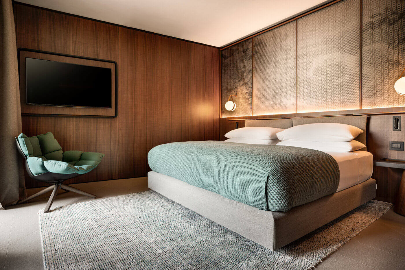 il Sereno Lago di Como, Europe — Modern luxury bedroom with walnut wood walls, sage linen bedding, pendant lighting, and contemporary green chair overlooking minimalist interior design.