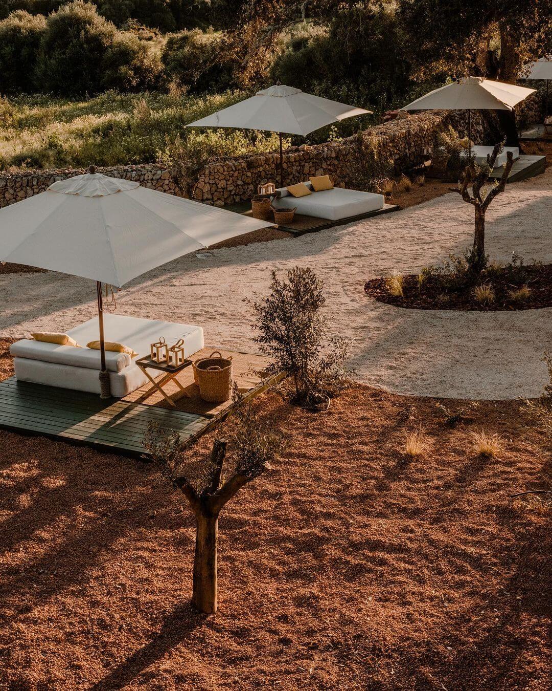 Amagatay, Balearic Islands — Terracotta courtyard with linen umbrellas, daybed seating, olive trees, and stone walls in Mediterranean minimalist design.