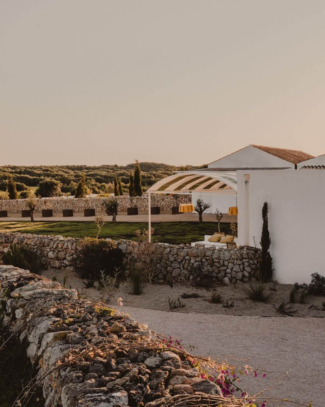 Amagatay, Balearic Islands — Minimalist white architecture with striped pergola, stone walls, and manicured grounds overlooking Mediterranean landscape at golden hour.