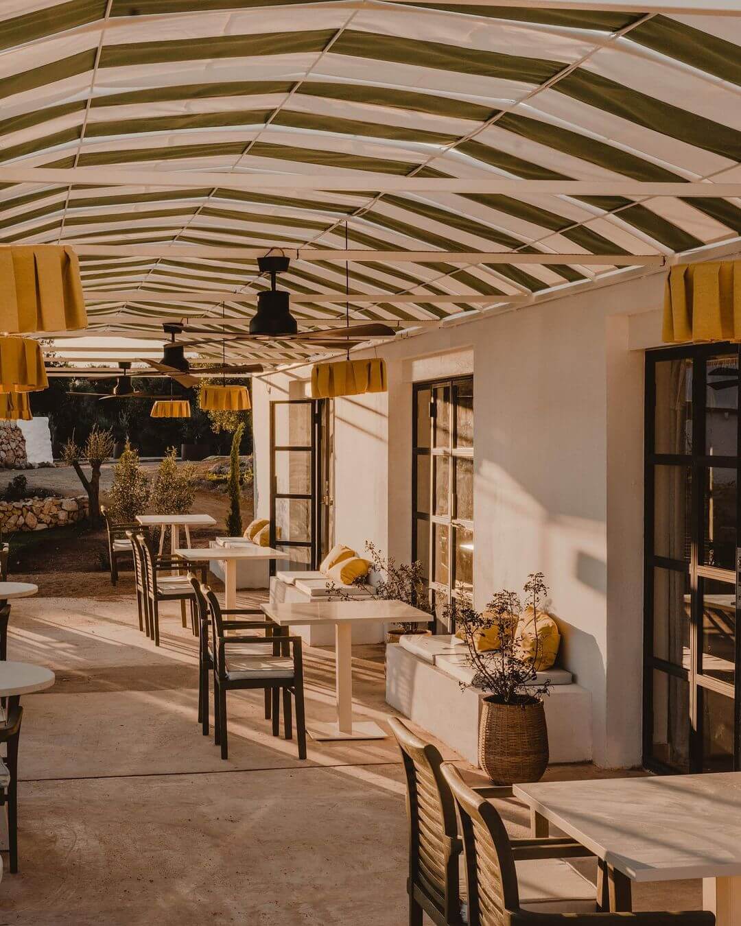 Amagatay, Balearic Islands — Covered terrace with striped white canvas roof, natural wood tables, black chairs, yellow pendant lights, and pale stone walls flooded with afternoon sun.