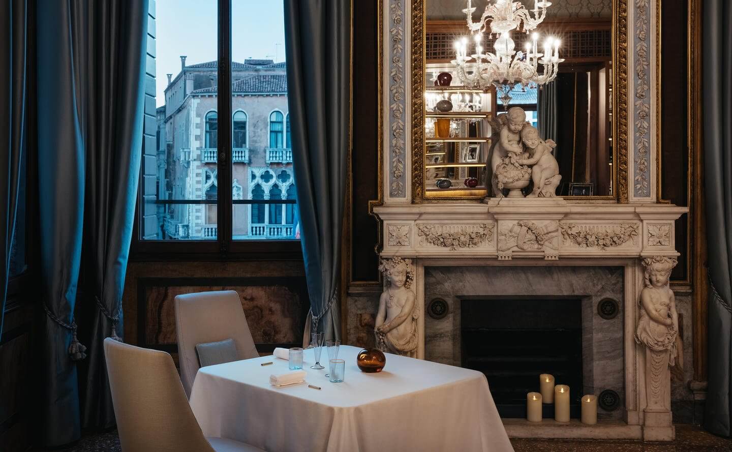 Cook the Lagoon at Aman, Venice, Italy — Neoclassical fireplace with marble mantelpiece, gilt-framed mirror, crystal chandelier, overlooking Grand Canal through draped windows.