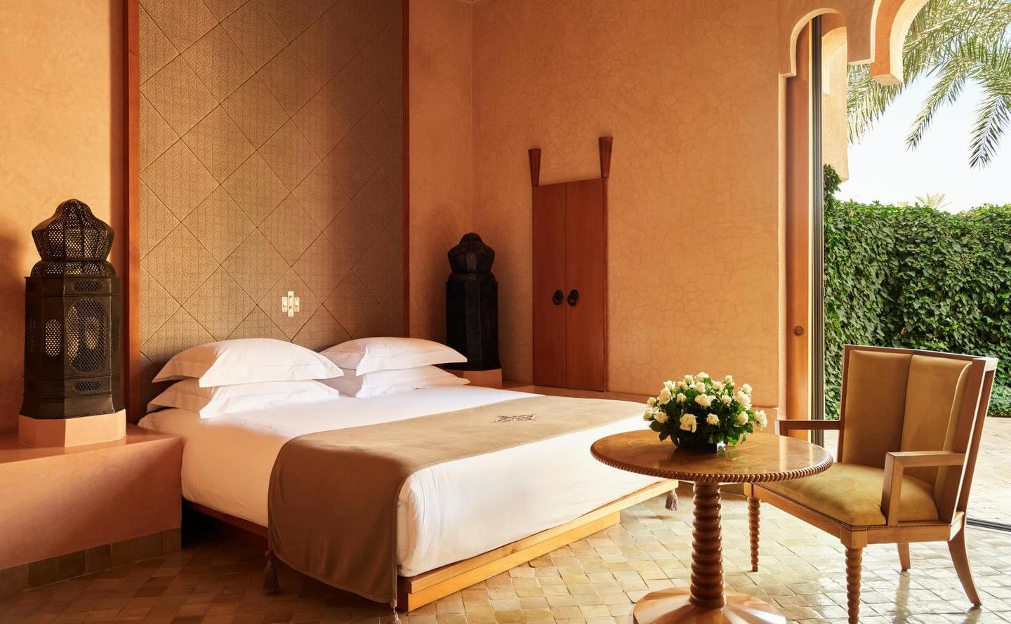 Amanjena, Africa — Ochre bedroom with carved wood furnishings, white linens, Moroccan lantern, and garden views through arched colonnade.