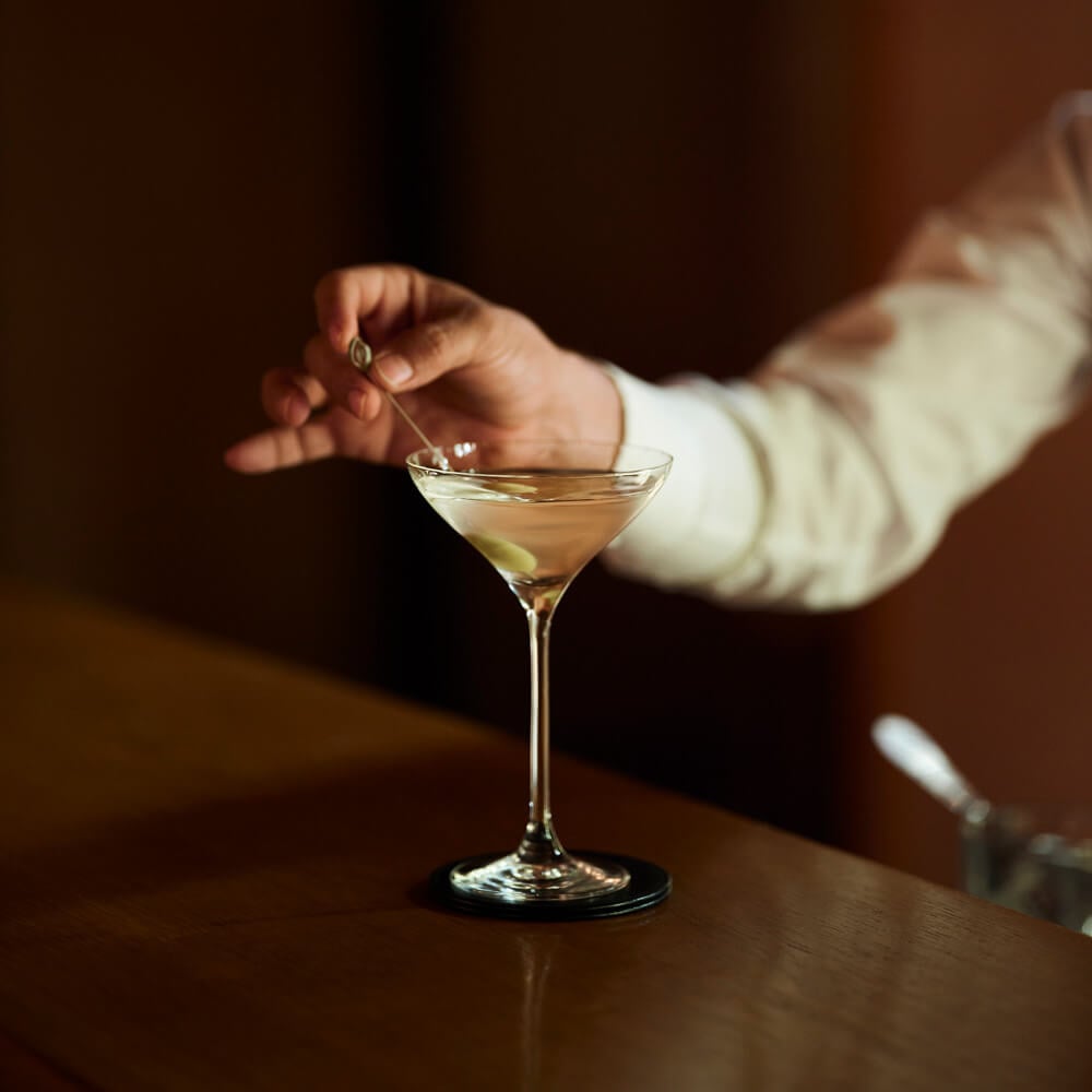 Amanjena, Africa — Bartender stirring dry martini with olive in coupe glass, cream linen sleeve against warm brown interior lighting.
