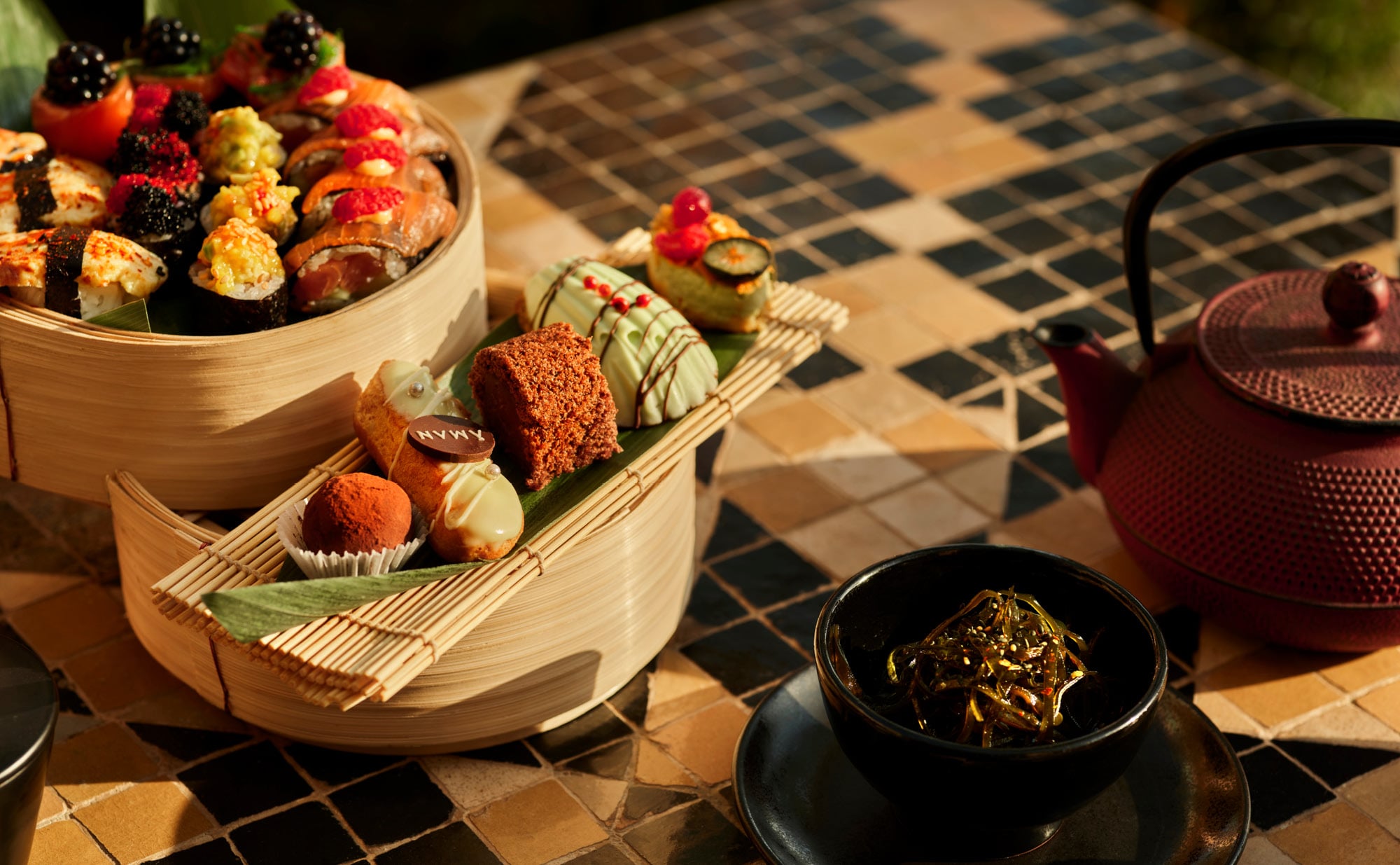 Amanjena, Africa — Tiered bamboo steamer baskets with artisanal chocolates and fresh berries beside cast-iron teapot on geometric tile floor, afternoon sunlight.