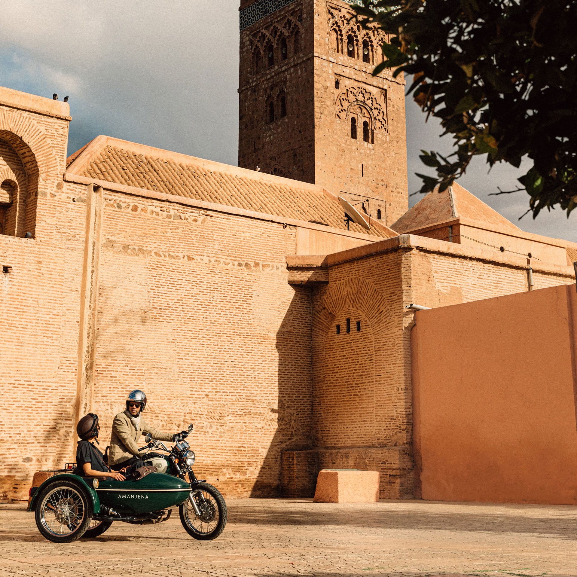 Marrakech by Motorbike, Morocco — sidecar motorcycle parked in ochre-walled medina courtyard with minaret tower and traditional Islamic architecture.