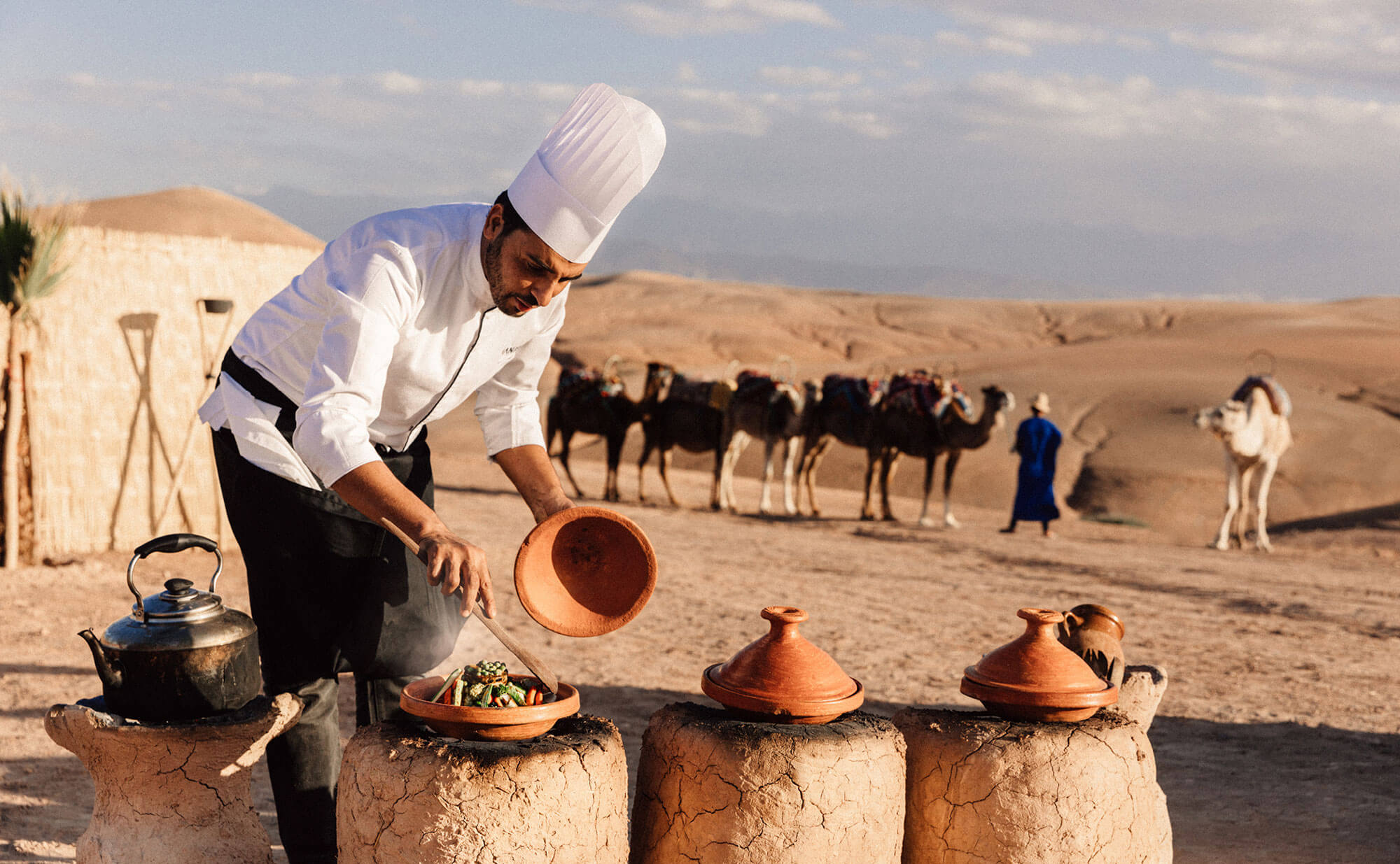 Amanjena, Africa — Chef in white chef's hat pouring preserved tagine over terracotta cookware in desert camp with camel caravan and sand dunes.