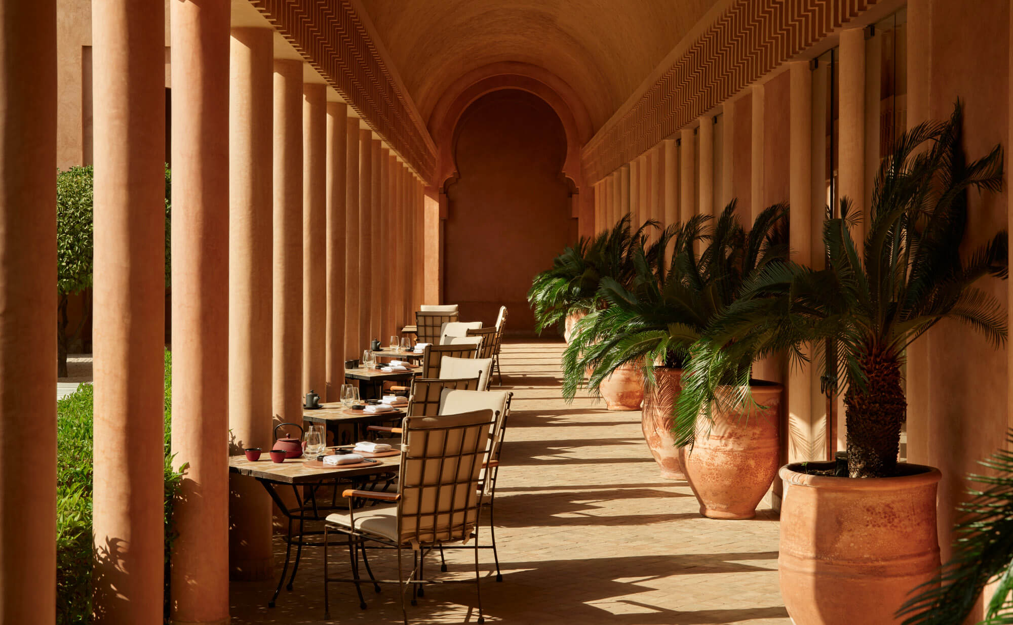 Amanjena, Marrakech, Morocco — Terracotta colonnade with arched ceiling, dining tables, potted palms, and dappled sunlight casting geometric shadows on stone floor.