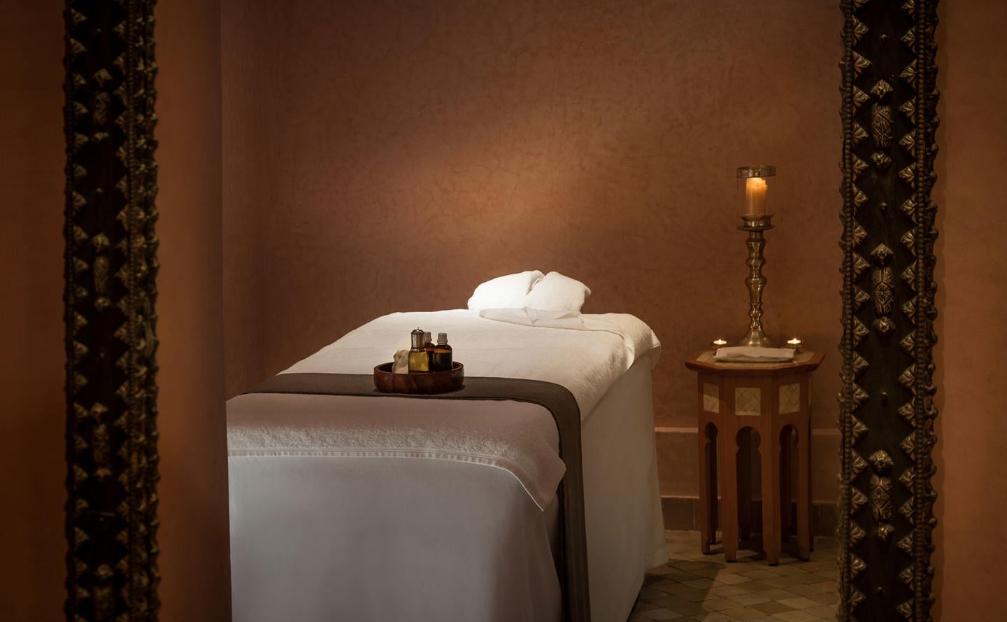 Amanjena Spa, Africa — Minimalist massage bed with white linens, brass candleholder, and decorative bottles in warm-toned spa room with ornate fabric panels.