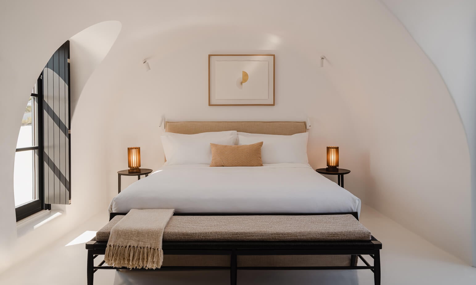 Nobu Santorini, Greece — Minimalist bedroom with curved whitewashed walls, natural linen bedding, wooden headboard, arched window with black shutters, and warm pendant lighting.