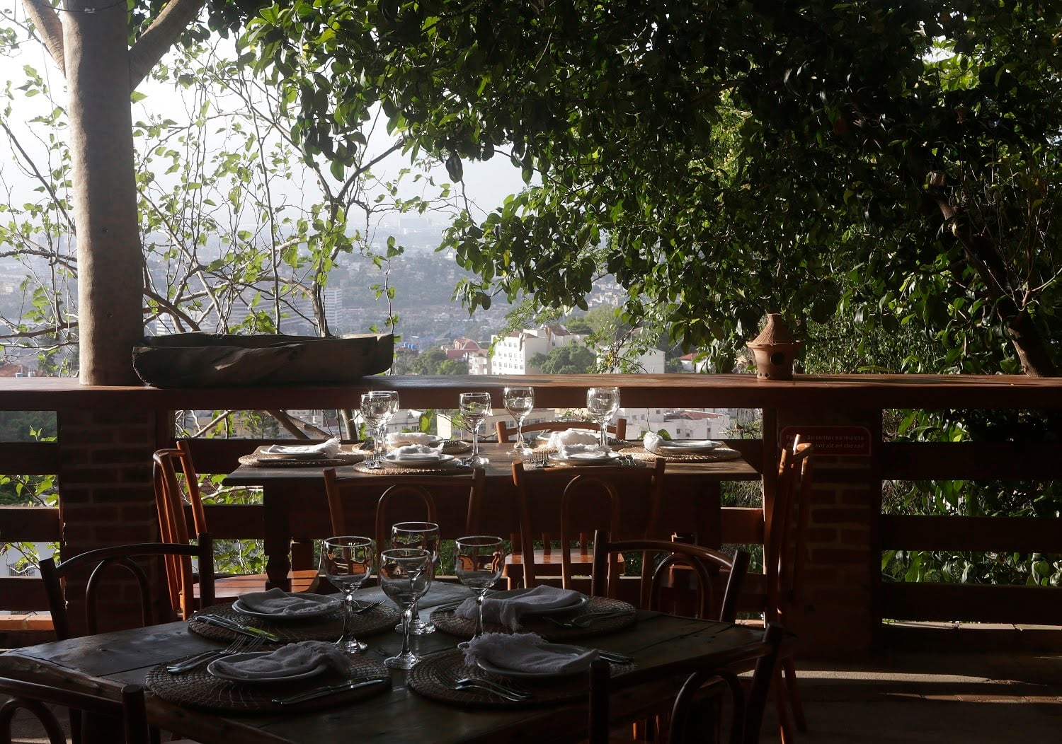 Aprazível, South America — Elevated dining terrace with dark wood tables, leaf-draped views over misty hillside city, natural light through leafy canopy, minimalist place settings.