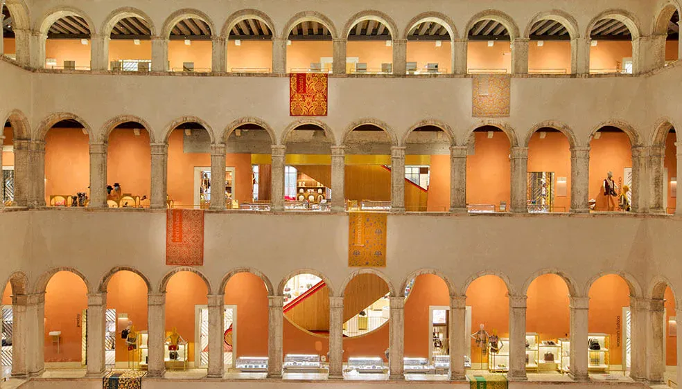 T Fondaco dei Tedeschi by DFS, Venice, Italy — Renaissance palazzo courtyard with three tiers of symmetrical arched colonnades, warm terracotta walls, and soft golden interior lighting.