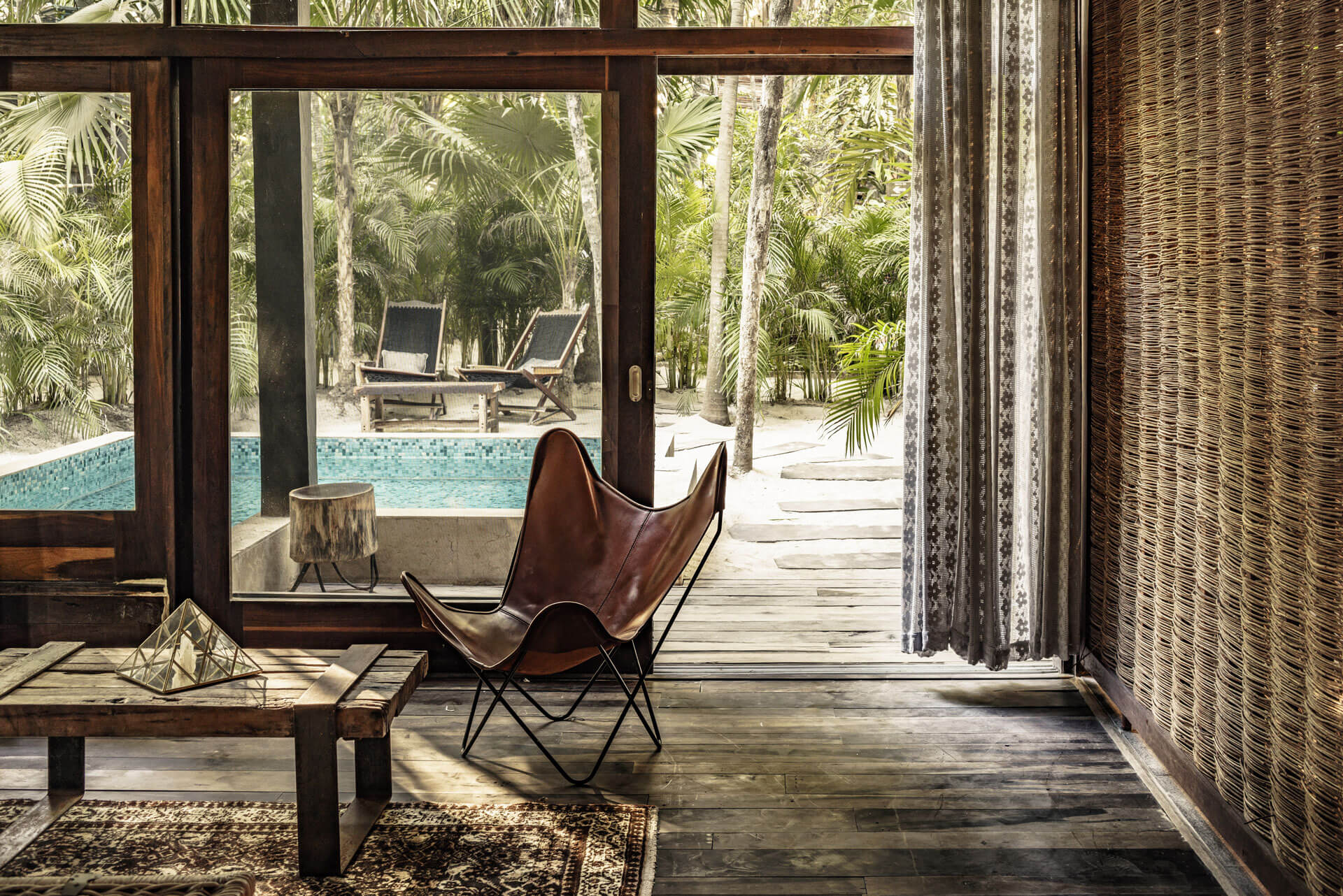 Be Tulum, Mexico — Modernist leather butterfly chair on wooden deck overlooking infinity pool, palm forest, and loungers in tropical resort interior.