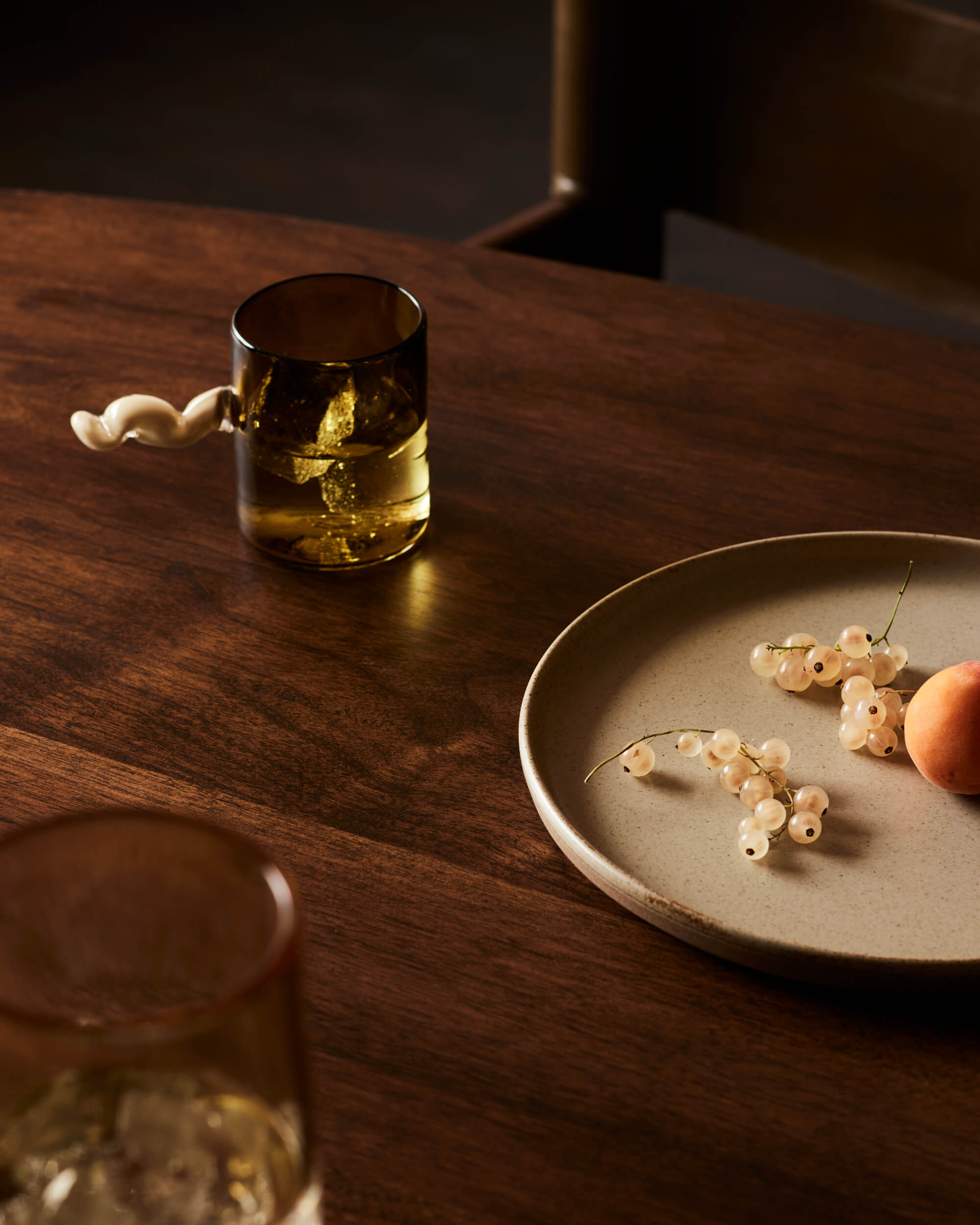 Arjé Home, North America — Wooden table set with amber glass, white plate holding white currants and apricot, warm natural light casting shadows, minimalist luxury dining aesthetic.