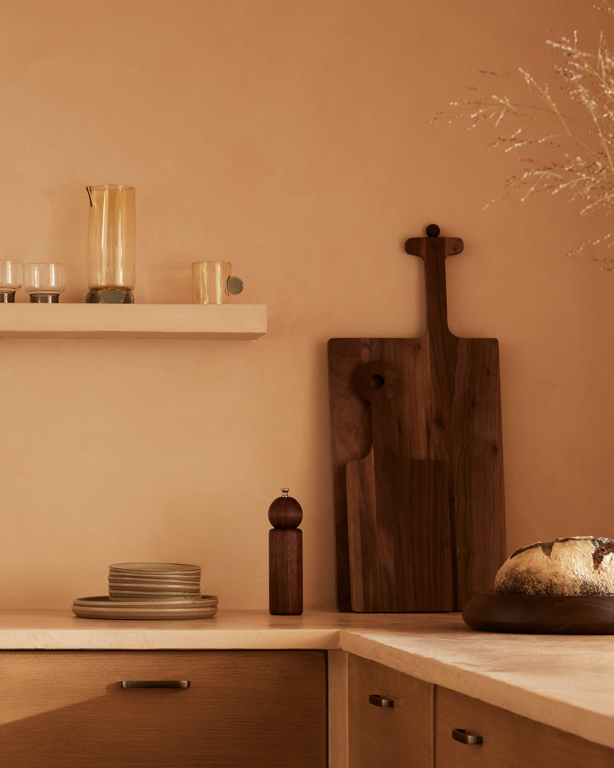Arjé Home, North America — Minimalist kitchen counter with warm terracotta walls, wooden cutting boards, ceramic tableware, and glass vessels on floating shelf.