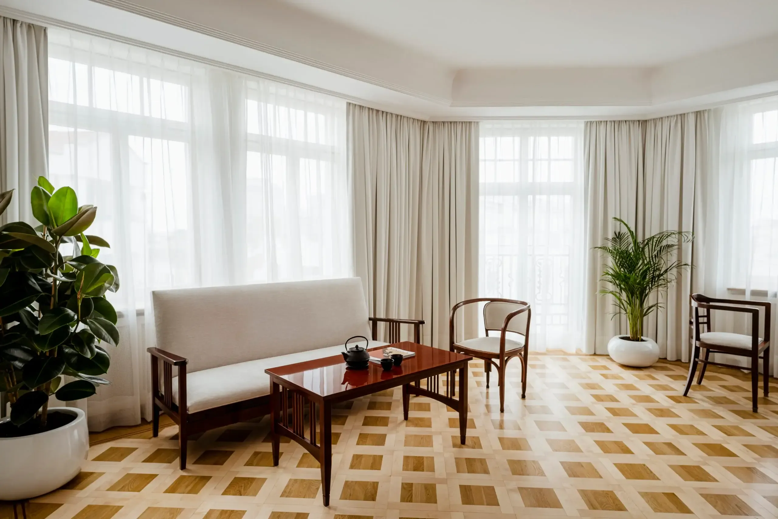 Nobu Warsaw, Poland — Minimalist hotel suite with mid-century wooden furniture, cream sofa, geometric parquet floor, floor-to-ceiling curtains, and abundant natural light from curved windows.