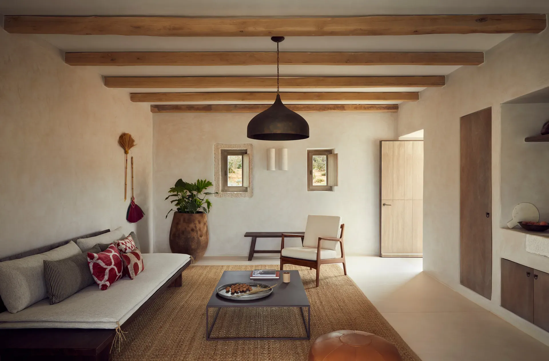 The Rooster, Europe — Minimalist villa living room with exposed wooden beams, black pendant light, cream linen sofa, mid-century chair, terracotta plant vessel, and natural daylight.