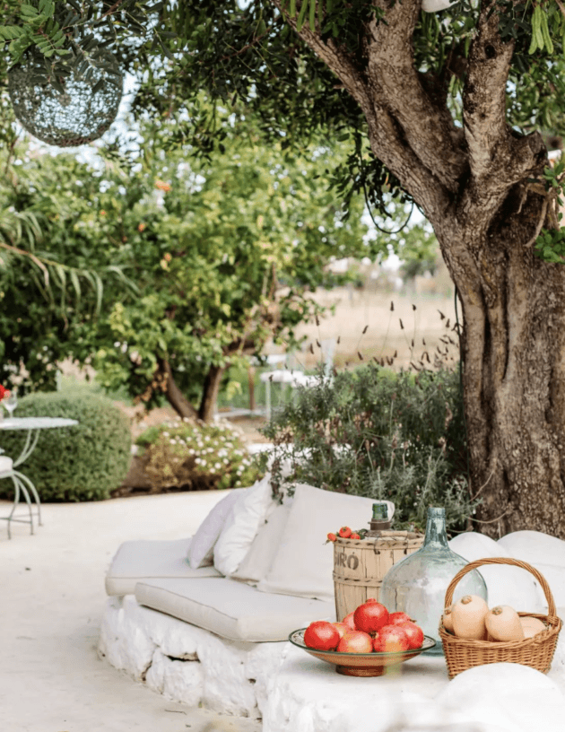 Aubergine, Balearic Islands — Rustic outdoor lounge beneath ancient olive tree with cream daybed, terracotta pottery, fresh produce, and Mediterranean garden view.