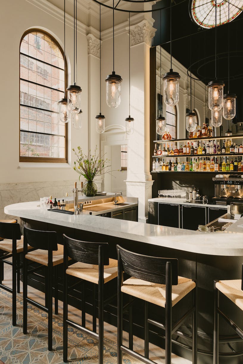 August Bar, Europe — Curved white marble counter with black wooden stools, pendant glass lighting, mirrored back bar, arched window, modern luxury hospitality design.