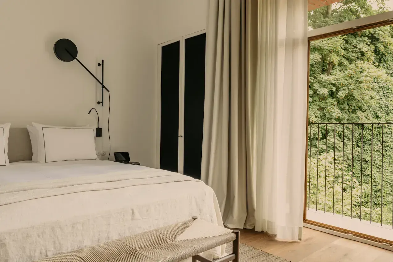 August, Europe — Minimalist bedroom with white linens, black pendant lamp, cream curtains, and glass doors opening to verdant landscape.