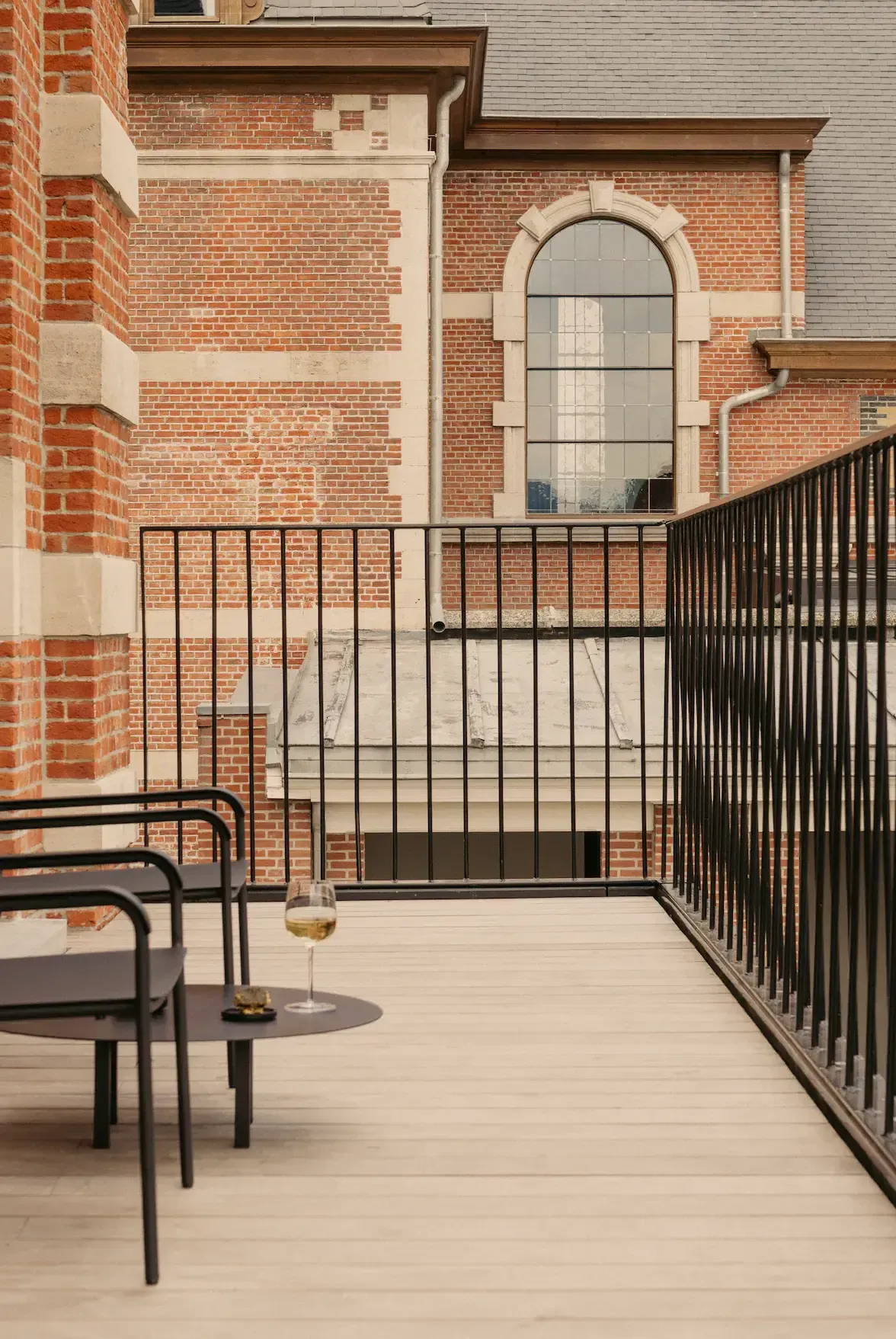 August, Europe — Rooftop terrace with brick architecture, arched window, black metal railings, light wood decking, and midcentury modern seating overlooking urban courtyard.