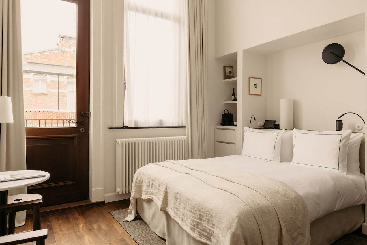 August Hotel, Europe — Minimalist bedroom with cream bedding, wooden herringbone floors, brass radiator, floor-to-ceiling windows, and built-in shelving with curated objects.