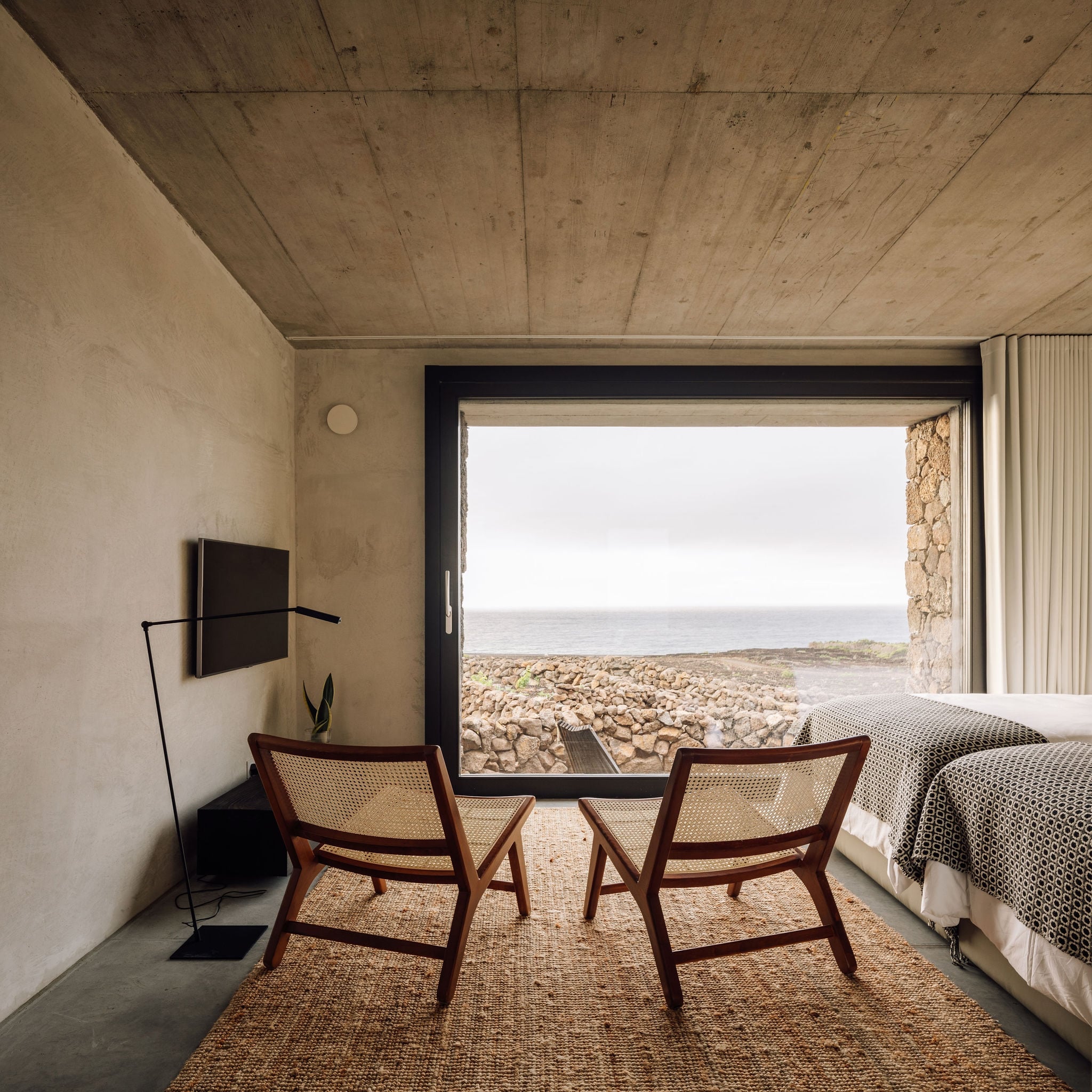 Azores Wine Company, Europe — Minimalist concrete bedroom with rattan chairs, floor-to-ceiling window framing rocky coastline and ocean horizon.