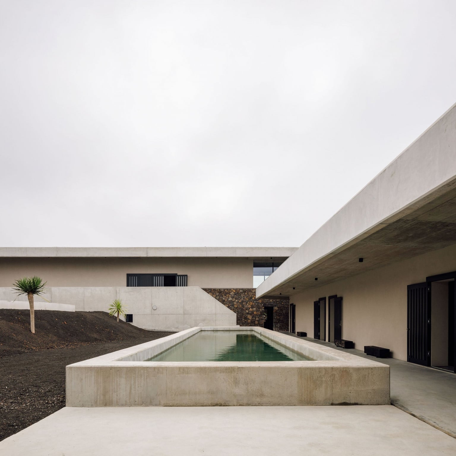 Azores Wine Company, Europe — Minimalist concrete courtyard with rectangular pool, modern architecture, volcanic stone accent wall, and overcast sky.