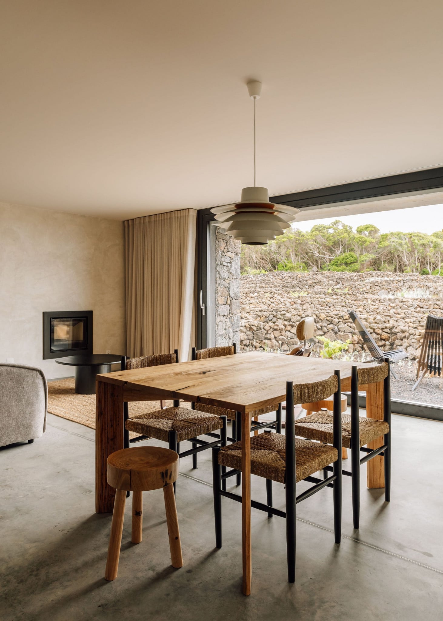 Azores Wine Company, Europe — Dining room with timber table, woven chairs, pendant light, stone fireplace, and panoramic windows overlooking rocky landscape.