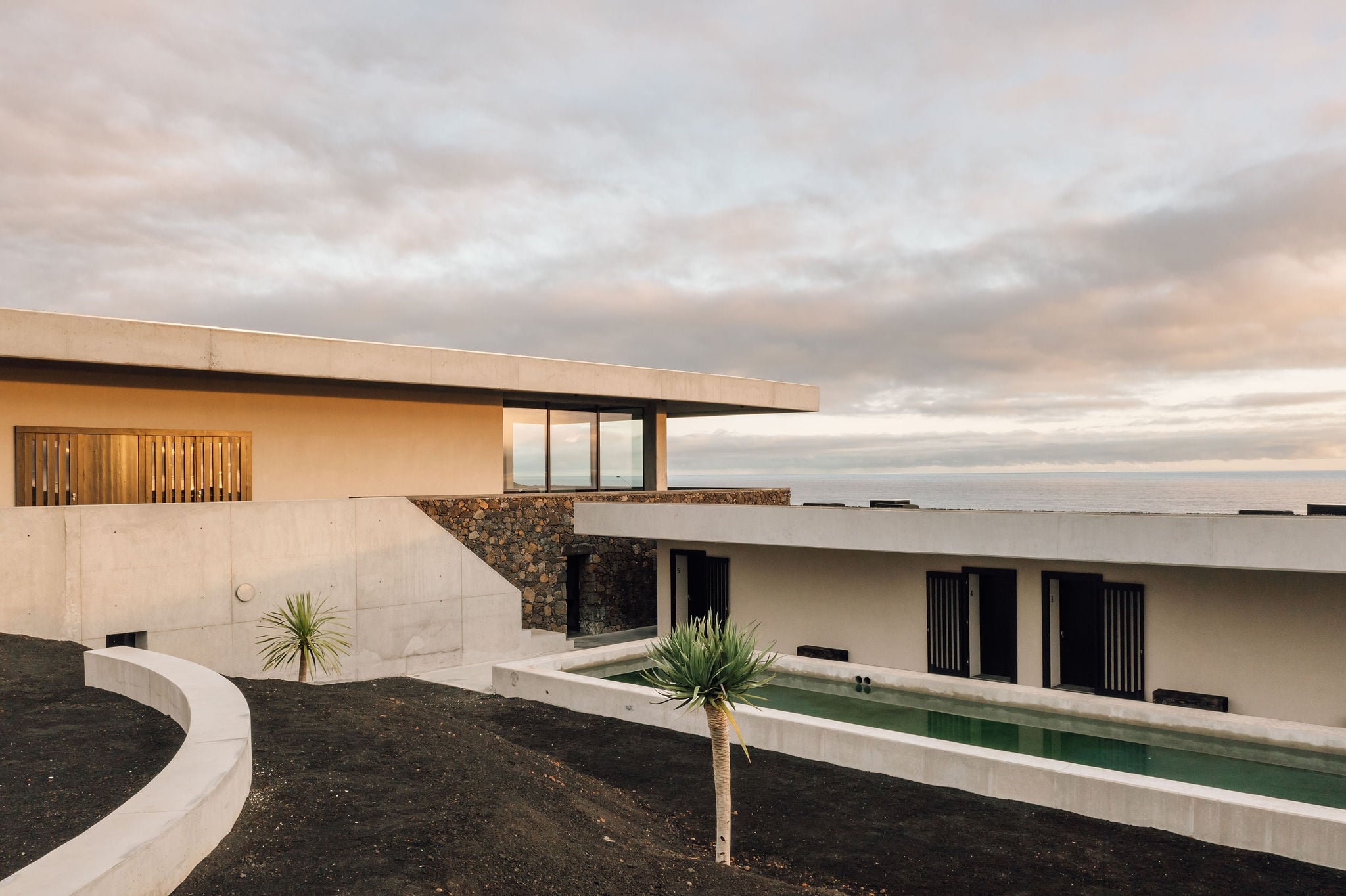 Azores Wine Company, Europe — Modernist concrete villa with volcanic stone accent, minimalist landscape, reflecting pool at dusk under clouded sky.