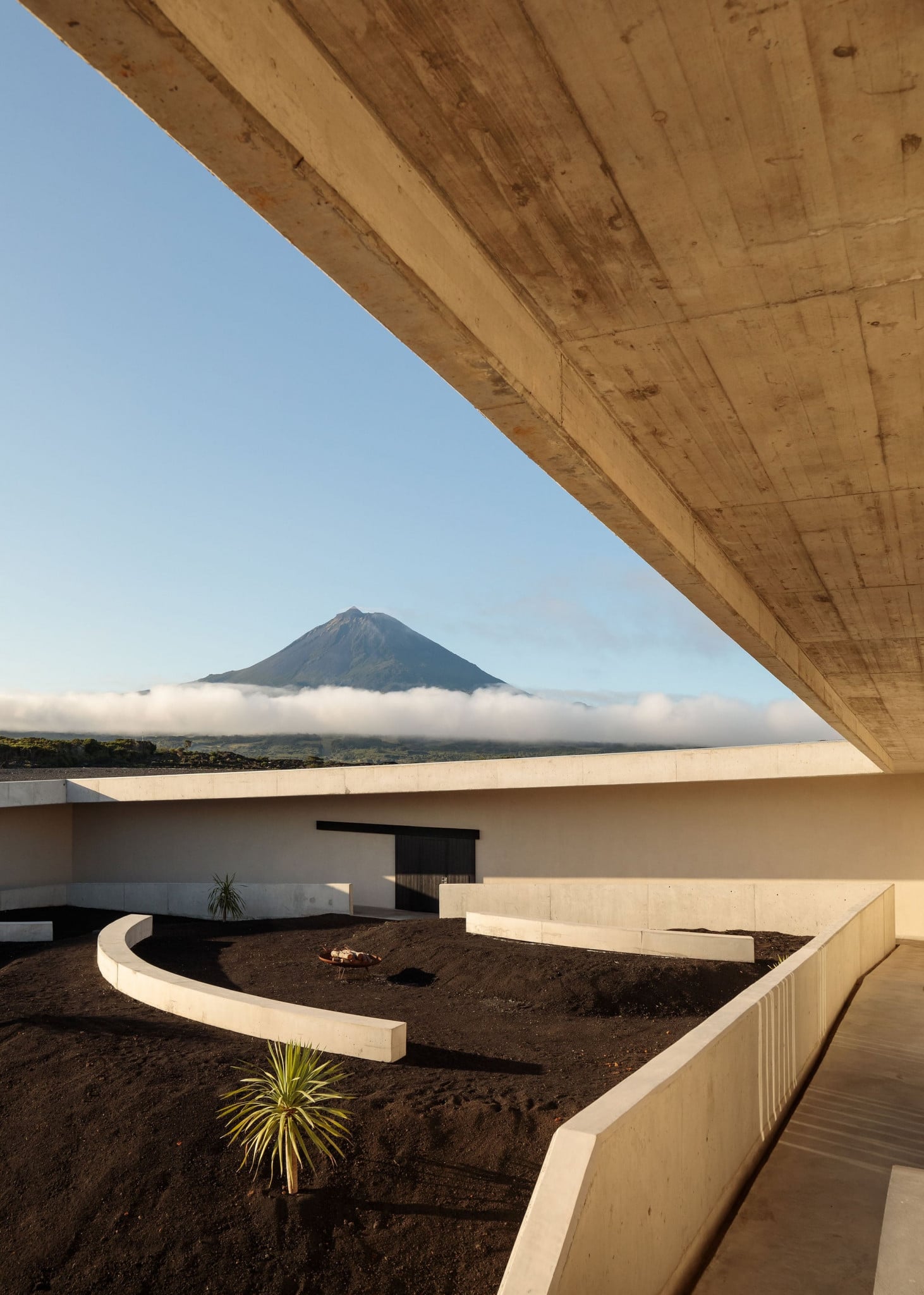 Azores Wine Company, Europe — Concrete interior courtyard framing volcanic mountain under mist, minimalist architecture with dark soil landscaping and wooden overhang.