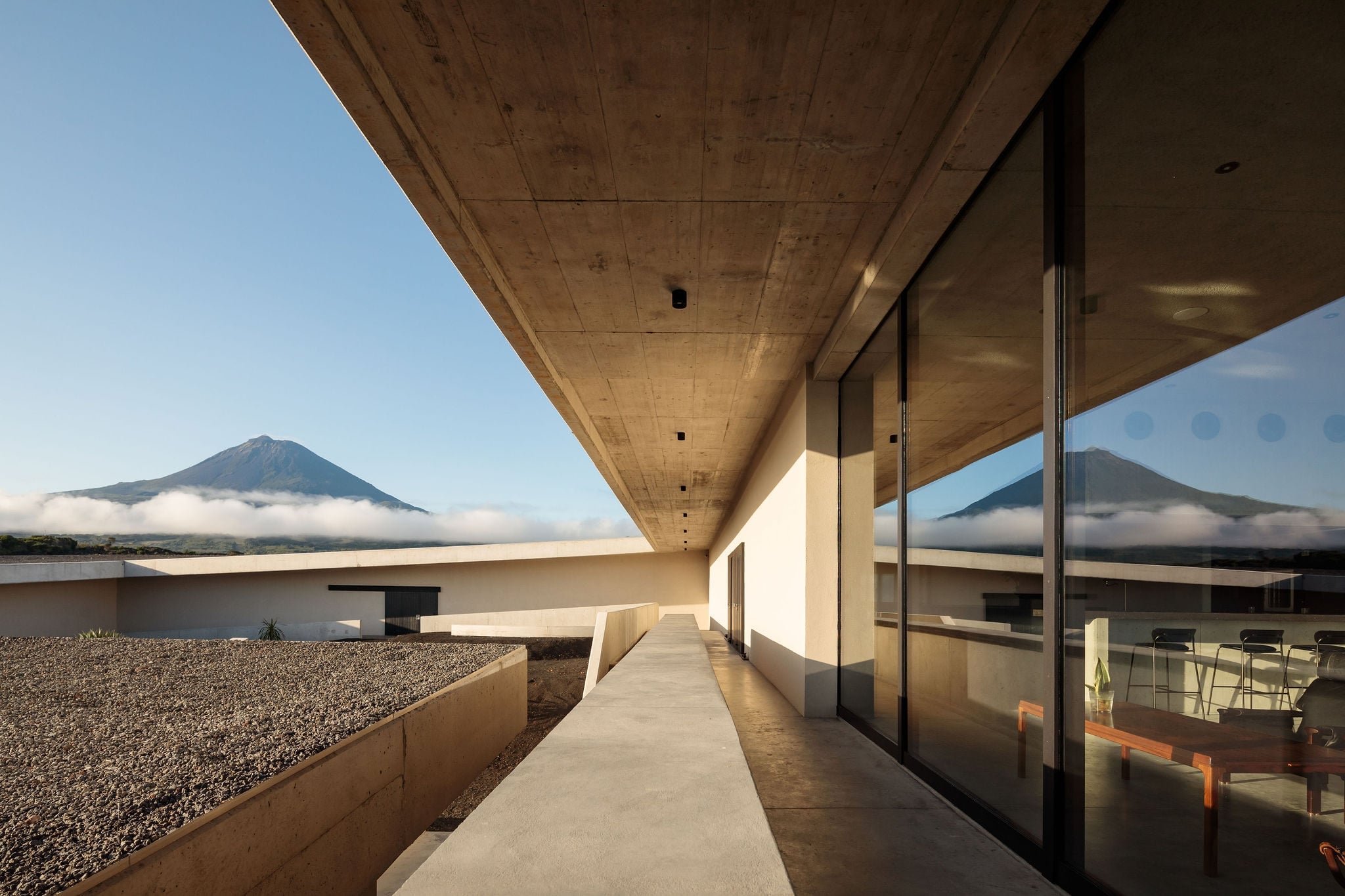 Azores Wine Company, Europe — Modernist concrete terrace with floor-to-ceiling windows overlooking volcanic peaks, minimalist aesthetic with gravel landscaping and natural light.
