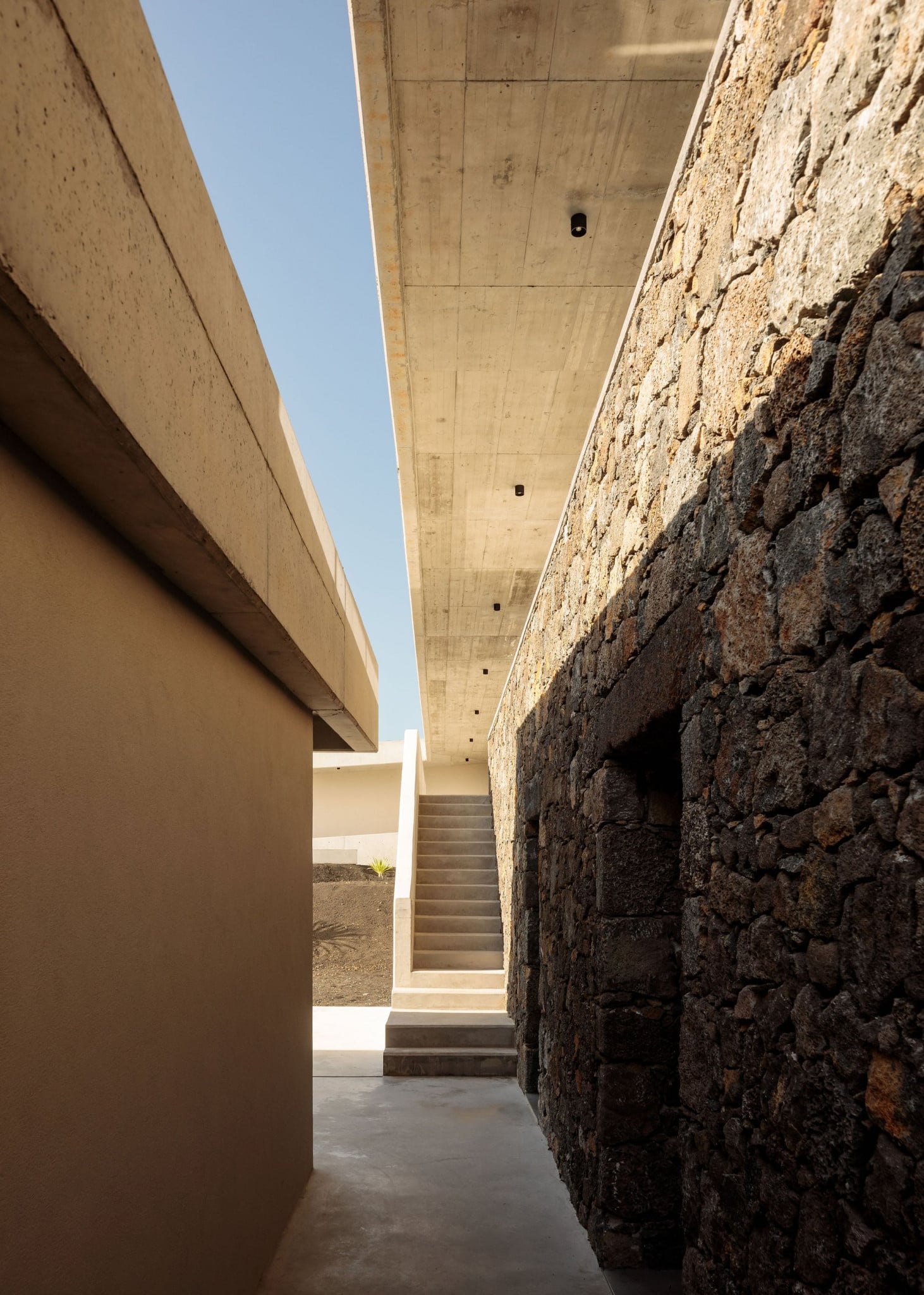 Azores Wine Company, Europe — Narrow corridor with raw stone wall, concrete beam ceiling, and minimalist staircase flooded with natural light.