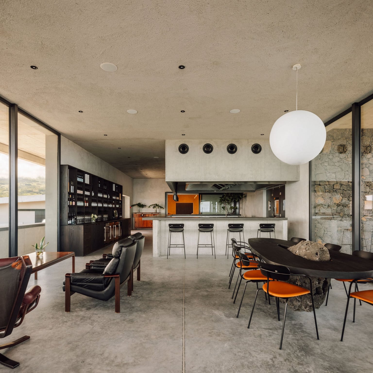 Azores Wine Company, Europe — Modern hospitality bar with concrete ceiling, black bar stools, orange chairs, stone accent wall, and spherical pendant light overlooking vineyard views.