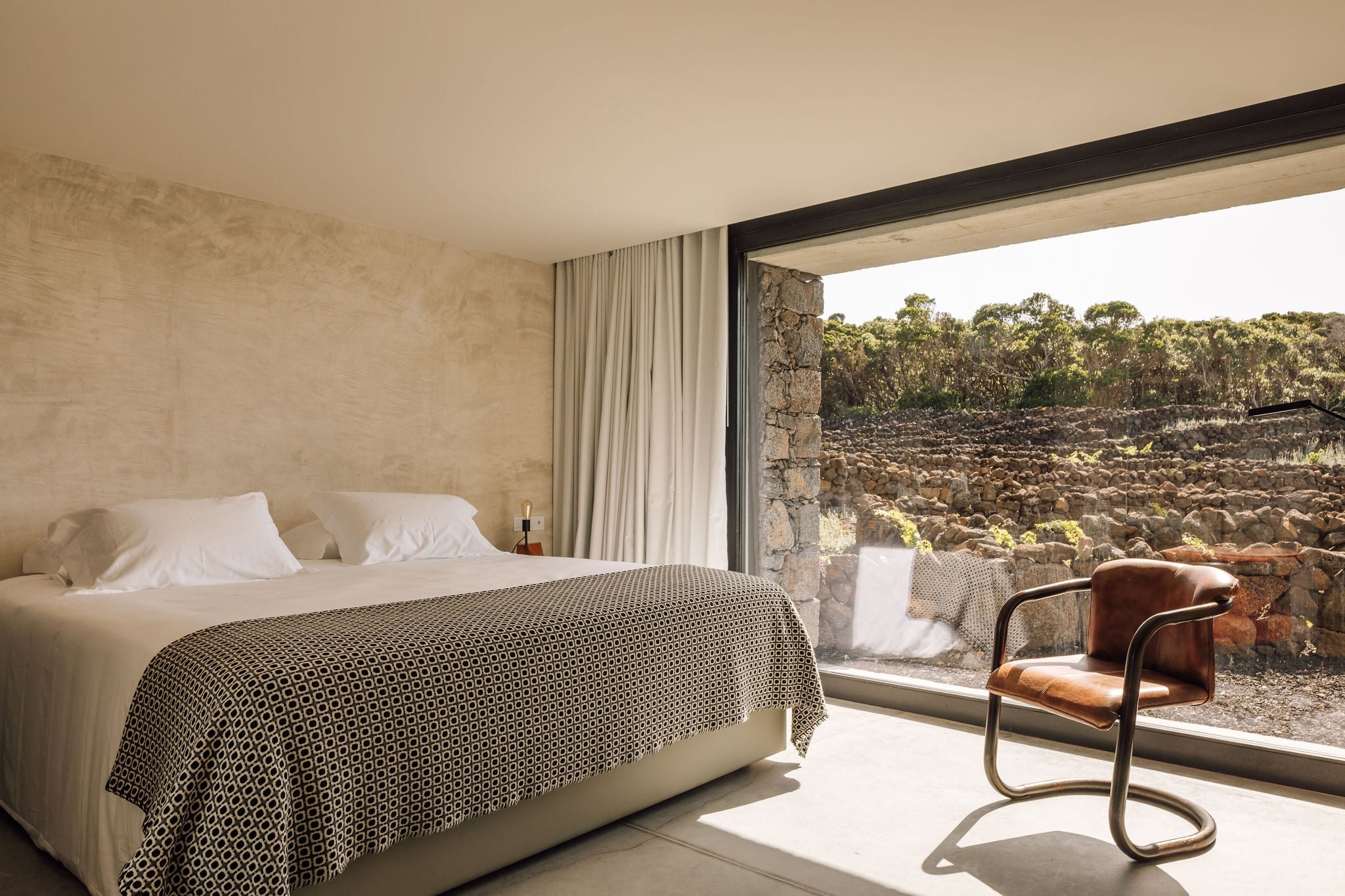 Azores Wine Company, Europe — Minimalist bedroom with stone terrace overlooking vineyard landscape, natural light, leather chair, geometric bedcover.