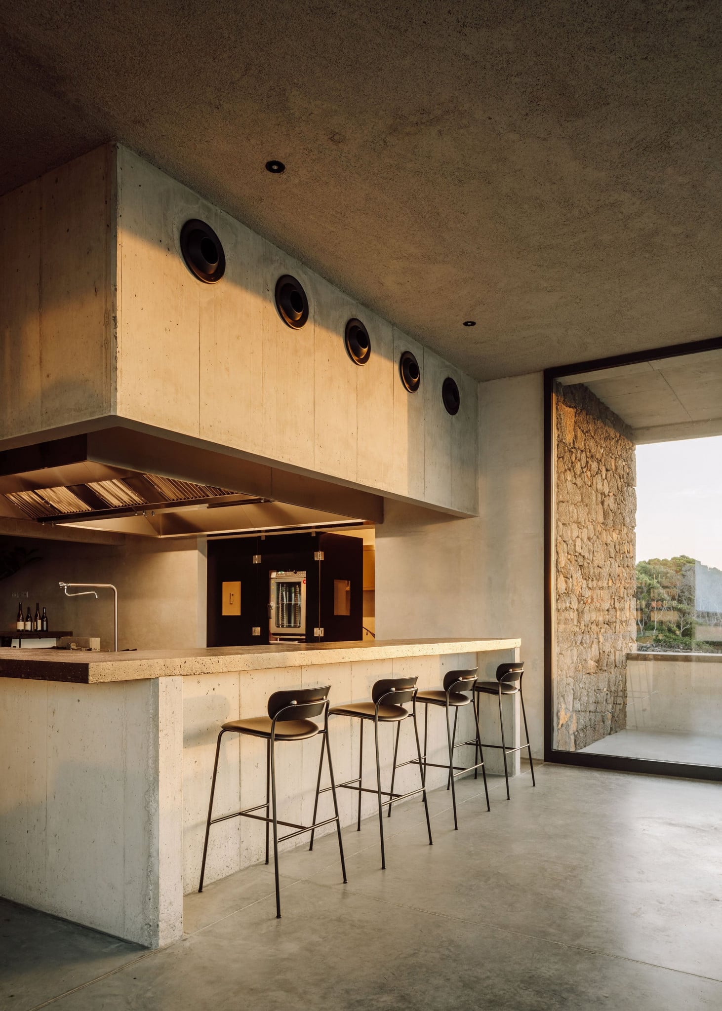 Azores Wine Company, Europe — Concrete kitchen with light wood cabinetry, bar seating, and stone accent wall overlooking landscape.